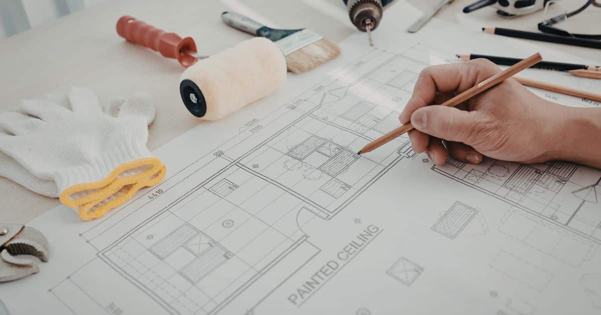 A close-up view of a person’s hands using a pencil to review and mark a detailed architectural floor plan. The plan is spread across a workspace and surrounded by essential renovation tools like a paint roller, brush, and work gloves