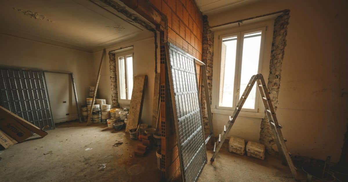 An interior view of a home undergoing a major remodel, featuring exposed brick walls, metal framing for pocket doors, a stepladder, buckets of plaster, and natural light coming through two large windows.