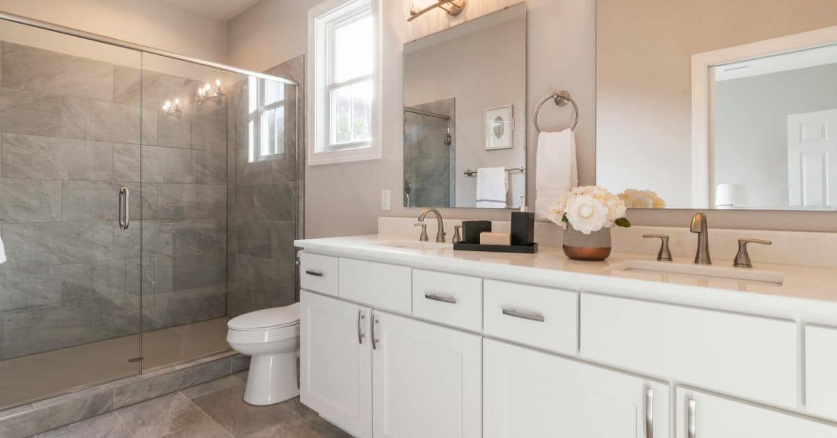 Creating a Spa-Like Experience with Bath Remodeling A clean, contemporary bathroom featuring a white double vanity, large mirrors, a white toilet, and a glass-enclosed walk-in shower with grey stone tiling.