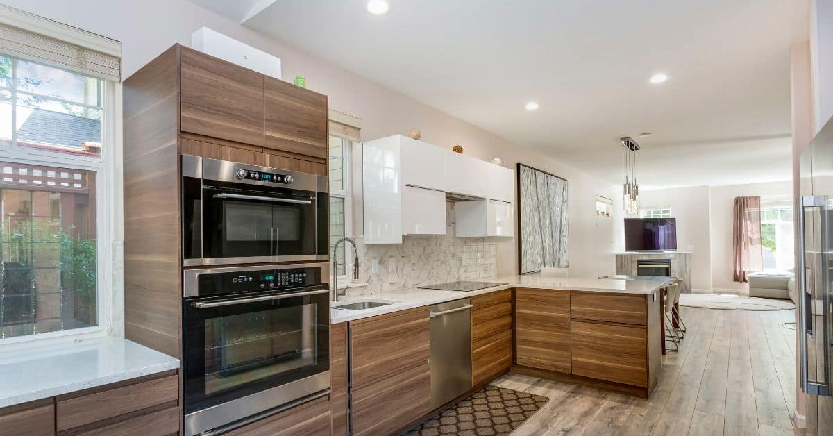 Functional Innovation in Modern Kitchen Design A bright, modern kitchen with wood-grain cabinetry, integrated stainless steel ovens, white countertops, and an open view into the living area with a fireplace.