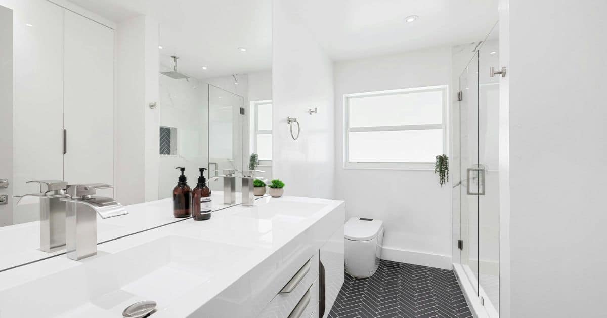 Minimalist Master Bathroom Design A clean, minimalist white bathroom featuring a large frameless glass shower, a wide vanity with double sinks, and dark herringbone floor tiles.